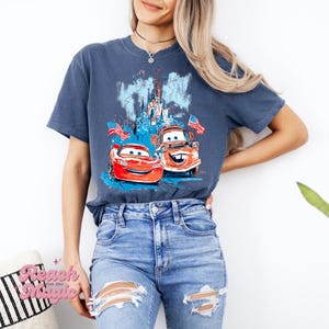 May include: A navy blue t-shirt with a graphic of two cartoon cars, American flags, and a castle. The cars are red and brown. The text "Reach for Magic" is on a pillow in the lower left corner.