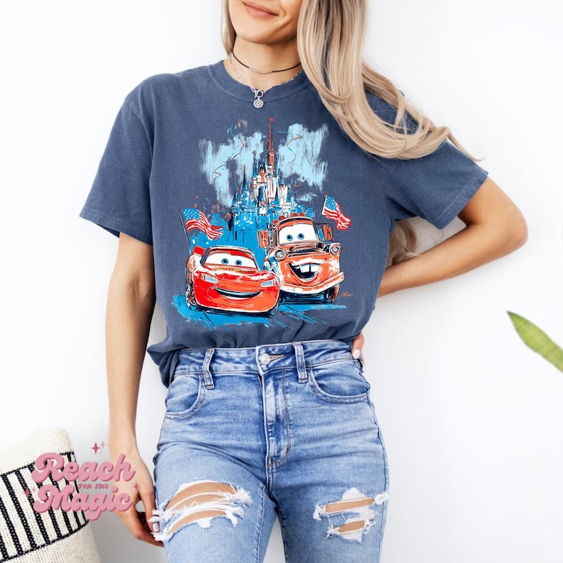 May include: A navy blue t-shirt with a graphic of two cartoon cars, American flags, and a castle. The cars are red and brown. The text "Reach for Magic" is on a pillow in the lower left corner.