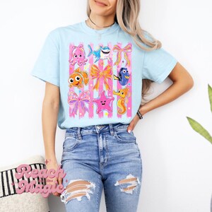 Könnte beinhalten: Ein hellblaues T-Shirt mit einem Grafikdruck verschiedener Meerestiere, darunter ein Oktopus, ein Hai, ein Clownfisch, ein Doktorfisch und ein Seepferdchen, durchsetzt mit rosa Schleifen und vertikalen rosa Streifen auf hellblauem Grund.