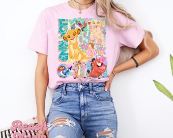 May include: Pink t-shirt featuring a colorful graphic of characters from The Lion King, including Simba, Timon, Pumbaa, and Zazu. The word "KING" is displayed vertically on the left side of the graphic. The shirt is made of a soft material.