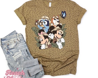 May include: A tan-colored t-shirt with a leopard print pattern. The shirt features a graphic of Mickey and Minnie Mouse, and Bluey and Bingo from Bluey, dressed in safari outfits. A pair of distressed blue jeans are folded next to the shirt.