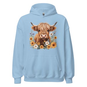 May include: Light blue hoodie featuring a watercolor illustration of a Highland cow surrounded by sunflowers and wildflowers. The cow has brown fur and large horns. The hoodie has a drawstring hood and a front pocket.