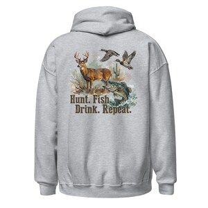 May include: A light gray hoodie featuring a graphic of a deer, fish, and ducks with the text "Hunt. Fish. Drink. Repeat." The design includes a deer, a fish, and two ducks in flight, with a nature scene.