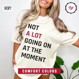 May include: Ivory-colored t-shirt with the text "NOT A LOT GOING ON AT THE MOMENT" in black and red. The shirt also has the words "COMFORT COLORS" in a red banner. A logo is visible in the upper right corner.