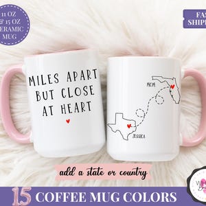 May include: Two white ceramic mugs with pink handles and interiors. One mug reads "MILES APART BUT CLOSE AT HEART" with a red heart. The other mug features a map with states and the text "MOM" and "JESSICA".