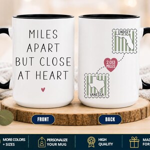 May include: A white ceramic mug with a black handle and rim. The front features the black text MILES APART BUT CLOSE AT HEART with a small pink heart. The back shows two postage stamp graphics with maps of Florida and Texas, connected by a dashed line and a pink heart labeled 2.013 MILES.