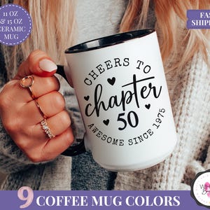 May include: White ceramic mug with black interior and handle. The mug features the text "Cheers to chapter 50 Awesome Since 1975" in black script. The mug is available in 11 oz and 15 oz sizes. The image also shows a hand holding the mug.