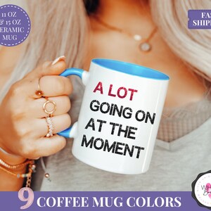 Puede incluir: Taza de caf&eacute; de cer&aacute;mica blanca con interior y asa azules. La taza presenta el texto "A LOT GOING ON AT THE MOMENT" en negro y rojo. Disponible en 9 colores y en tama&ntilde;os de 325 ml y 444 ml.