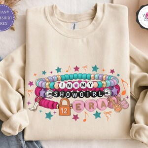 Flaylay of an ivory Gildan youth sweatshirt with an In My Showgirl Era friendship bracelet design in pink and teal taylor hoodie, kids taylor hoodie, youth sweatshirt, girls sweatshirt, toddler sweatshirt, youth crewneck, taylor apparel, taylor girls clothes, showgirl hoodie, kids showgirl shirt, youth showgirl tee, girls taylor shirt, showgirl costume
