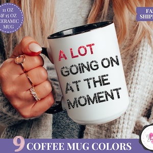 Puede incluir: Taza de cer&aacute;mica blanca con interior y asa negros, con la inscripci&oacute;n "A LOT GOING ON AT THE MOMENT" en negro y rojo. Disponible en tama&ntilde;os de 325 ml y 444 ml. La imagen tambi&eacute;n muestra el texto "9 COFFEE MUG COLORS".