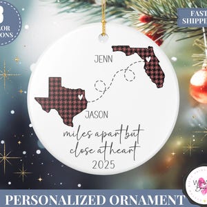 May include: A white ceramic personalized ornament with a gold string for hanging. The ornament features the states of Texas and Florida with a heart design, connected by a dotted line. Text includes names, "miles apart but close at heart," and the year 2025.