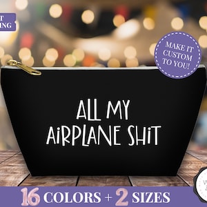 May include: Black zippered pouch with the text "ALL MY AIRPLANE SHIT" in white. The bag has a gold zipper pull and is on a wooden surface. Additional text includes "MAKE IT CUSTOM TO YOU!" and "16 COLORS + 2 SIZES".