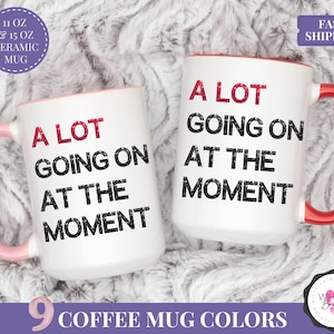 Puede incluir: Dos tazas de cer&aacute;mica blancas con la frase "A LOT GOING ON AT THE MOMENT" impresa en negro y rojo. Una taza tiene un asa y un interior rosa, la otra tiene un asa y un interior rojos. Las tazas est&aacute;n disponibles en 312 ml y 444 ml.