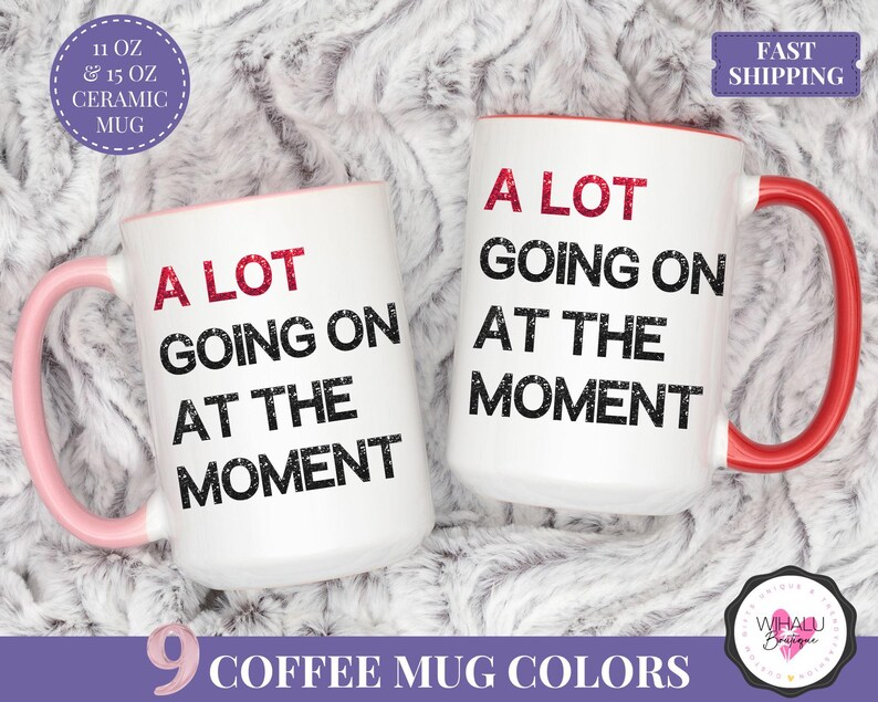 Puede incluir: Dos tazas de cer&aacute;mica blancas con la frase "A LOT GOING ON AT THE MOMENT" impresa en negro y rojo. Una taza tiene un asa y un interior rosa, la otra tiene un asa y un interior rojos. Las tazas est&aacute;n disponibles en 312 ml y 444 ml.