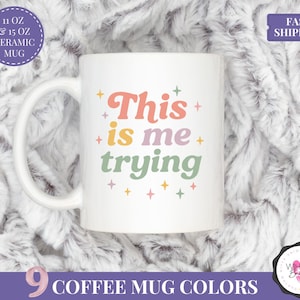 May include: White ceramic mug with the text "This is me trying" in a retro font. The mug is available in 11 oz and 15 oz sizes. The image also includes text indicating "9 Coffee Mug Colours" and "Fast Shipping".