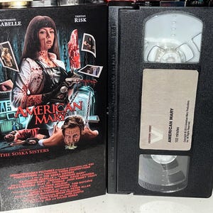 Pode incluir: Uma fita VHS preta e a sua capa de cartão para o filme American Mary. A capa mostra uma mulher com um avental manchado de sangue a segurar um bisturi, com o texto American Mary e The Soska Sisters. A fita tem uma etiqueta com o título.
