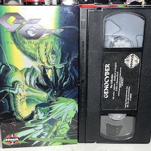 Pode incluir: Uma fita VHS Genocyber The Collection e a sua capa de cartão. A capa apresenta uma ilustração de estilo anime em verde e preto com o título Genocyber The Collection. A fita VHS preta tem uma etiqueta com o título e detalhes técnicos.