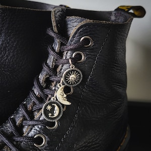 Może przedstawiać: Czarne skórzane buty z czarnymi sznurowadłami i srebrnymi zawieszkami w kształcie słońca, księżyca i półksiężyca z gwiazdą.