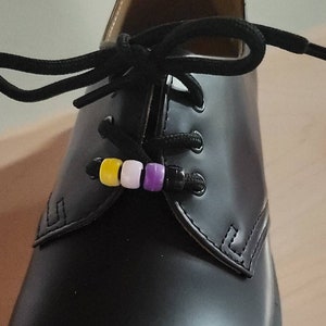 May include: Black leather shoe with black laces, featuring a non-binary pride flag bead charm. The charm includes yellow, white, and purple beads. The shoe has decorative stitching and a yellow sole.