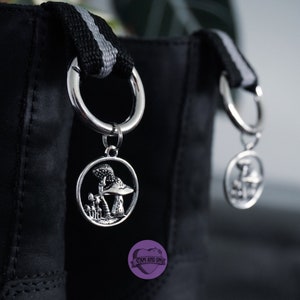 以下が含まれることがあります： 黒革のバッグに銀色のチャームが2つ付いている。それぞれのチャームは、細かなデザインが施されたキノコの形をしている。バッグには黒と白のストライプのストラップが付いている。