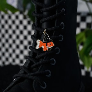 Puede incluir: Bota negra con cordones y un charm temático de Halloween. El charm presenta un pez naranja y blanco con un sombrero de bruja negro. El charm está sujeto a los cordones de la bota. El fondo está borroso, con un patrón de cuadros y vegetación.