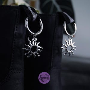 Può includere: Borsa in pelle nera con due ciondoli a forma di sole argentati attaccati alle cinghie. Le cinghie sono a righe bianche e nere.