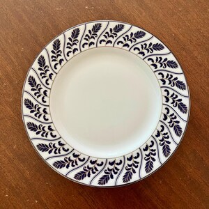 May include: A white ceramic plate with a navy blue leaf pattern around the rim and a gold trim. The plate has a simple, elegant design, suitable for serving food. The plate is round.