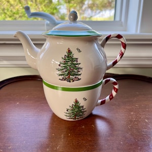 May include: A white ceramic teapot and teacup set with a Christmas tree design. The teapot has a green rim and a candy cane-striped handle. The teacup also features a green rim and a matching Christmas tree design. The set is on a wooden surface.