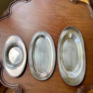 May include: Three oval silver-toned metal serving dishes of varying sizes arranged in a row. Each dish features a smooth, reflective interior basin and a textured, decorative border along the outer rim, showcasing a metallic finish.