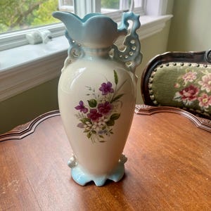 May include: A cream-colored ceramic pitcher featuring a floral bouquet of purple and white flowers on the front. The pitcher has a light blue scalloped rim, a decorative ornate handle with cut-out details, and a light blue scalloped base.