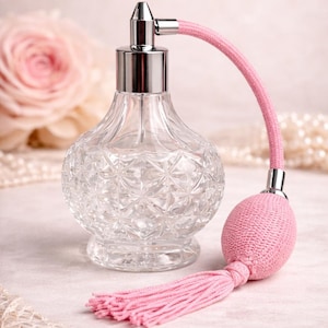 Puede incluir: Un frasco de perfume de vidrio transparente y facetado con una tapa de metal plateado y un atomizador rosa. El atomizador rosa tiene una pera y una borla a juego. El frasco está sobre una superficie rosa claro.
