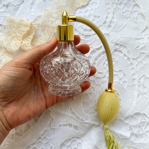 Puede incluir: Un frasco de perfume de vidrio transparente con una tapa dorada y un borla amarilla. El frasco tiene un diseño texturizado y se sostiene en una mano.
