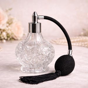 May include: A clear glass perfume bottle with a silver top and a black bulb atomizer. The bottle has a cut-glass design. The atomizer has a black woven tube and a black tassel. The background includes flowers and pearls.