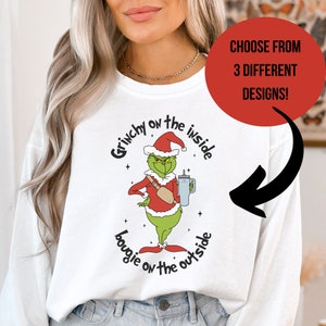 May include: White long-sleeve sweatshirt with a green Grinch character wearing a Santa hat and holding a cup. The text on the shirt reads "Grinchy on the inside, bougie on the outside."