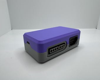 Puede incluir: Una consola de juegos de estilo retro en morado y gris. La consola tiene forma rectangular con puertos en la parte delantera y lateral. La parte superior es morada y la inferior gris.