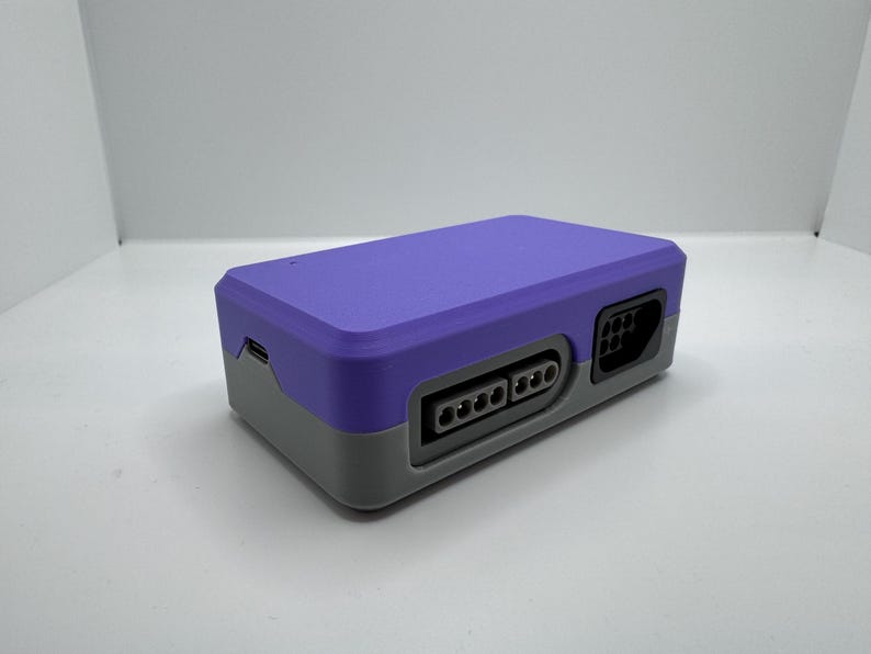 May include: A purple and gray retro-style game console. The console has a rectangular shape with ports on the front and side. The top is purple, and the bottom is gray.