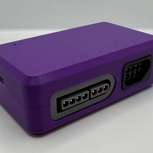 May include: A rectangular, purple electronic device with multiple ports. The device has a textured surface and a dark gray border around the ports. The ports include a USB port and several other connector ports.