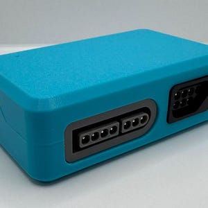 May include: A rectangular, teal-colored electronic device with multiple ports and connectors. The device has a USB port and several rectangular and circular ports. The device is likely a video game console or a related accessory.
