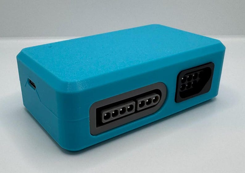 May include: A rectangular, teal-colored electronic device with multiple ports and connectors. The device has a USB port and several rectangular and circular ports. The device is likely a video game console or a related accessory.