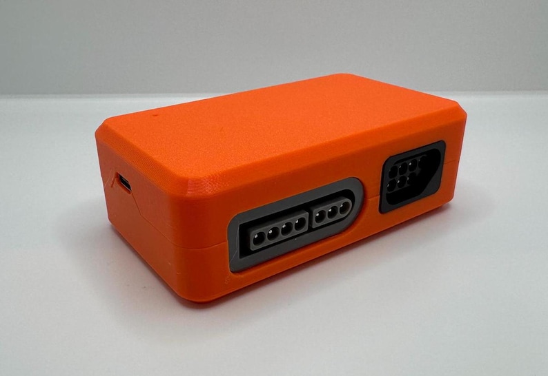 May include: Bright orange rectangular electronic device with multiple ports and connectors. The device has a textured surface and a sleek, modern design. It appears to be a console or adapter for gaming or other electronic applications.