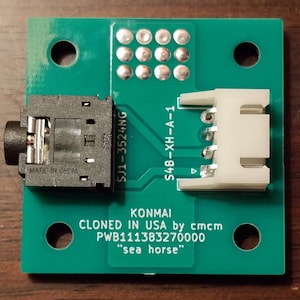 May include: A green circuit board with a black audio jack connector, a white connector, and text that reads "KONMAI CLONED IN USA by cmcm PWB111383270000 "sea horse""