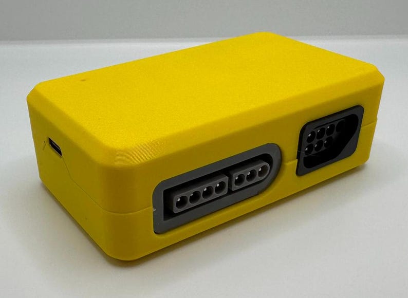 May include: A bright yellow rectangular electronic device with multiple ports. The device has a textured surface and a dark gray port panel with multiple pin connectors. A USB port is visible on the side.