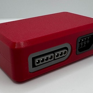 May include: A rectangular, red electronic device with multiple ports and connectors. The device has a textured surface and a gray border around the ports. It appears to be a retro gaming console accessory.