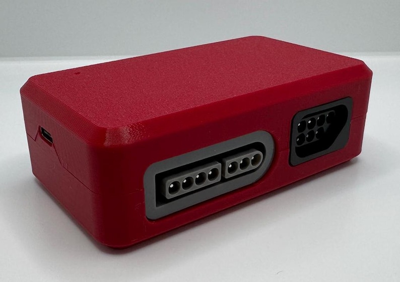 May include: A rectangular, red electronic device with multiple ports and connectors. The device has a textured surface and a gray border around the ports. It appears to be a retro gaming console accessory.