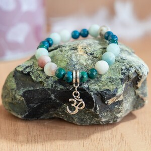 Könnte beinhalten: Ein Armband mit Perlen und einem silbernen Om-Anhänger. Das Armband besteht aus grünen, blauen und weißen Perlen.