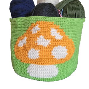 May include: A green crocheted basket with a mushroom design. The basket is filled with balls of yarn in various colors, including white, navy blue, and olive green.