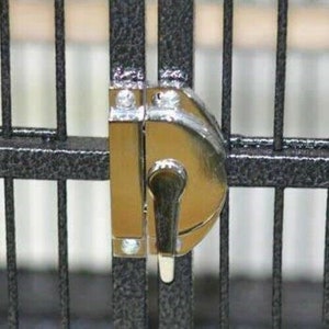 May include: A silver-toned metal latch mechanism secured to a black, textured metal cage door. The latch features a rounded, polished surface with a vertical handle that rotates to lock the two vertical bars of the cage together securely.