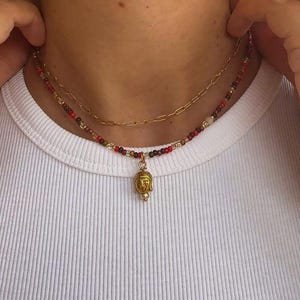 Könnte beinhalten: Ein mehrlagiges Halskettenset bestehend aus einer goldfarbenen Kette mit länglichen ovalen Gliedern und einer zweiten Kette mit kleinen, bunten Glasperlen in Rot-, Braun- und Cremetönen, ergänzt durch einen kleinen goldfarbenen Buddha-Anhänger.