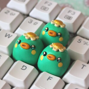 Op de afbeelding: Drie kleine, groene, vogelvormige figuurtjes met gele snavels en witte bloemen op hun koppen. De figuurtjes zitten op een wit toetsenbord.