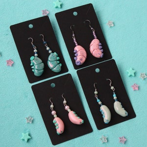 May include: Four pairs of handmade earrings displayed on black cards. Each pair features a unique design with a worm-like shape, adorned with beads and metallic hooks. The earrings come in various colors, including teal, pink, and light blue.